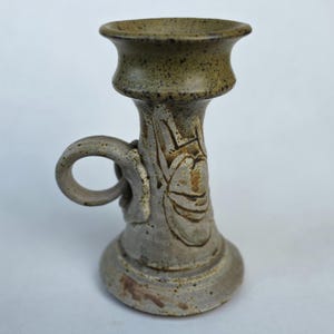 May include: A handcrafted ceramic candlestick holder with a handle. The candlestick is primarily gray with brown and green accents. It features a flared top and a detailed floral design on the body. The handle is a simple loop.