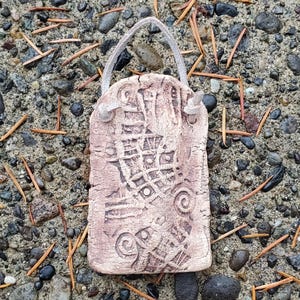 May include: A rectangular, light brown ceramic ornament with an abstract design and a loop for hanging. The surface has a textured appearance with carved patterns, including spirals and geometric shapes. The background consists of pebbles and pine needles.