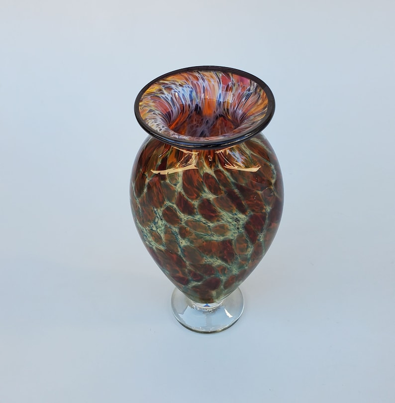 May include: A brown and green glass vase with a mottled pattern. The vase has a wide mouth and a narrow base. The vase is on a white background.