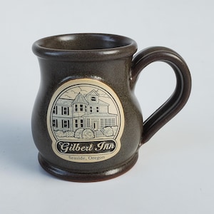 May include: A brown ceramic mug with a rounded body and a curved handle. The mug features a cream-colored oval emblem with a detailed illustration of a building and the text "Gilbert Inn Seaside, Oregon".