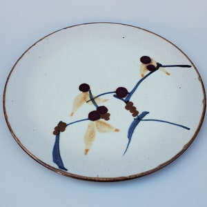 May include: A white ceramic plate with a brown rim features a hand-painted design of a stylized branch with brown and blue accents and yellow flowers.