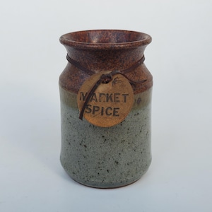 May include: A ceramic spice jar with a brown rim and a speckled grey-green body. A leather tag with the words "MARKET SPICE" hangs from a leather cord around the jar's neck. The jar has a rustic, handmade appearance.
