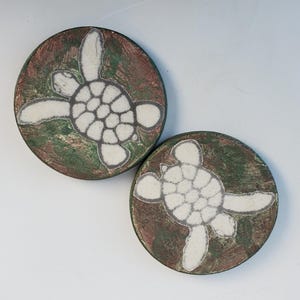 May include: Two round ceramic coasters with a green and brown marbled background. Each coaster features a white silhouette of a sea turtle.