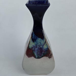 May include: A handcrafted ceramic vase with a unique shape and color scheme. The vase features a square top and a flared neck, transitioning to a faceted body. The colors include a deep navy blue, white, and a blend of red, green, and blue.