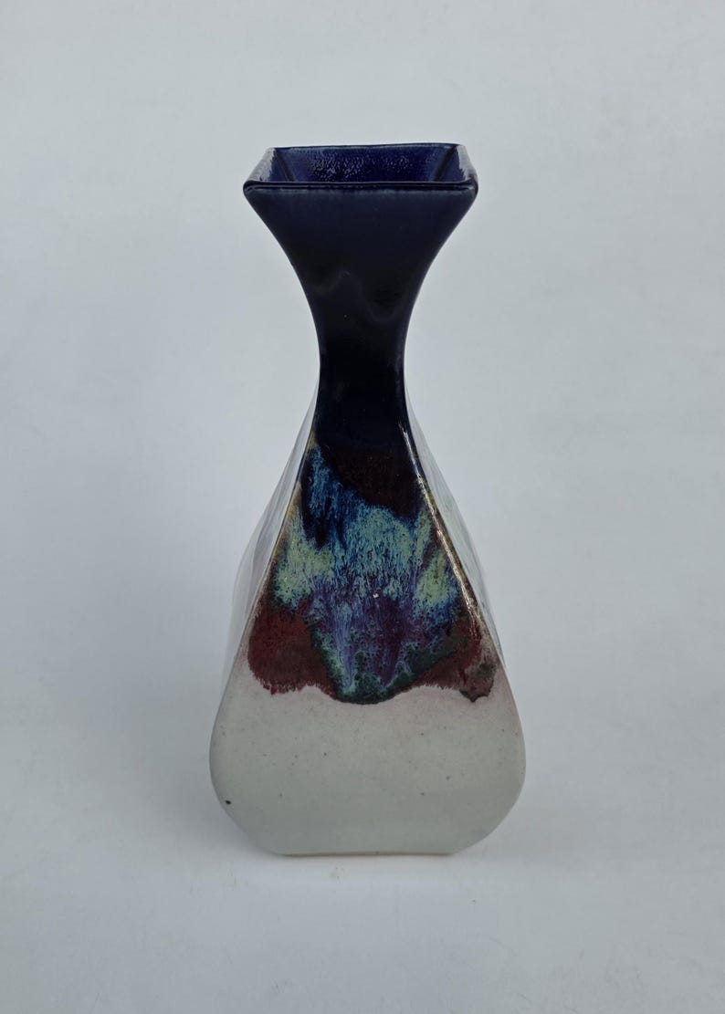 May include: A handcrafted ceramic vase with a unique shape and color scheme. The vase features a square top and a flared neck, transitioning to a faceted body. The colors include a deep navy blue, white, and a blend of red, green, and blue.