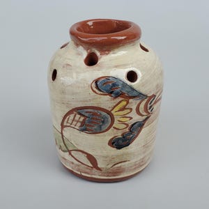 May include: A small, white ceramic vase with a brown rim and a floral design painted in blue, yellow, and green. The vase has several holes around the top for holding flowers.