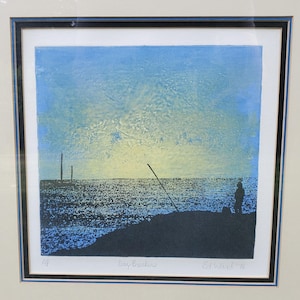May include: Framed print titled "Day Breakers" by Ed Ward '76. The artwork depicts a coastal scene with a silhouetted figure, a long pole, and two vertical structures against a blue and yellow sky. The print is framed in black.