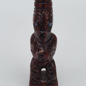 May include: A carved wooden tiki statue with a dark brown finish. The statue features intricate carvings, including facial details and patterns. The eyes are inlaid with blue stones. The tiki is in a seated position.