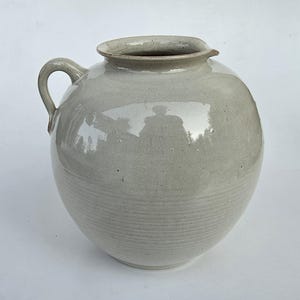 May include: A round, light gray ceramic jug with a glossy finish and a single side handle. The vessel features a wide, open mouth with a small spout detail and subtle horizontal ridges encircling the lower portion of the rounded body.