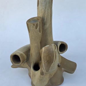May include: A beige ceramic vase with multiple cylindrical openings and a textured surface. The vase has a unique, organic shape with several protruding tubes and openings, suitable for floral arrangements or decorative display.