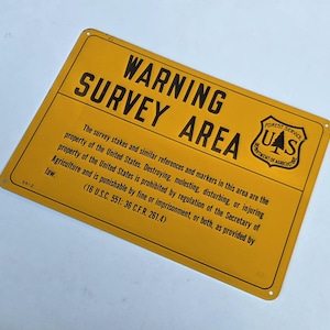May include: A yellow rectangular metal sign with black text. The sign reads "WARNING SURVEY AREA" and includes the U.S. Forest Service logo and legal text about survey markers.