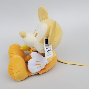 Puede incluir: Un juguete de peluche Mickey Mouse amarillo sentado en una superficie blanca. El juguete tiene una etiqueta pegada a su oreja derecha que dice "Disney Store" y "Seiko".