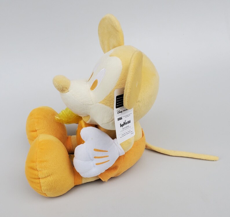Puede incluir: Un juguete de peluche Mickey Mouse amarillo sentado en una superficie blanca. El juguete tiene una etiqueta pegada a su oreja derecha que dice "Disney Store" y "Seiko".