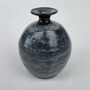 May include: A spherical ceramic vase with a narrow neck and a flared rim. The surface features a mottled glaze with deep black, charcoal gray, and subtle blue tones, creating a textured, layered appearance across the rounded body of the vessel.