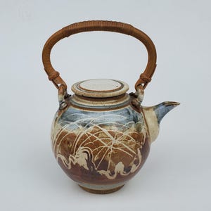May include: A ceramic teapot with a brown woven handle and lid. The body features a brown, blue, and white abstract design. The spout is blue and the lid is white.