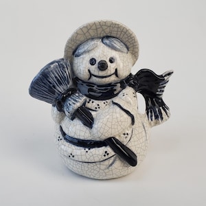 May include: A ceramic snowman figurine with a hat, scarf, and broom. The snowman is white with a crackle glaze and navy blue accents. The broom and scarf are navy blue. The snowman has a smiling face and is a winter holiday decoration.