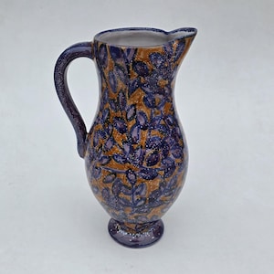 May include: A ceramic pitcher with a handle and spout, featuring a pattern of blue leaves on a tan background. The pitcher has a dark blue rim, handle, and base, with white speckles throughout the design. A decorative piece for the home.