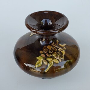 May include: A small brown ceramic vase with a wide opening and a floral design. The vase has a glossy finish and features a raised floral design in shades of brown, yellow, and green.