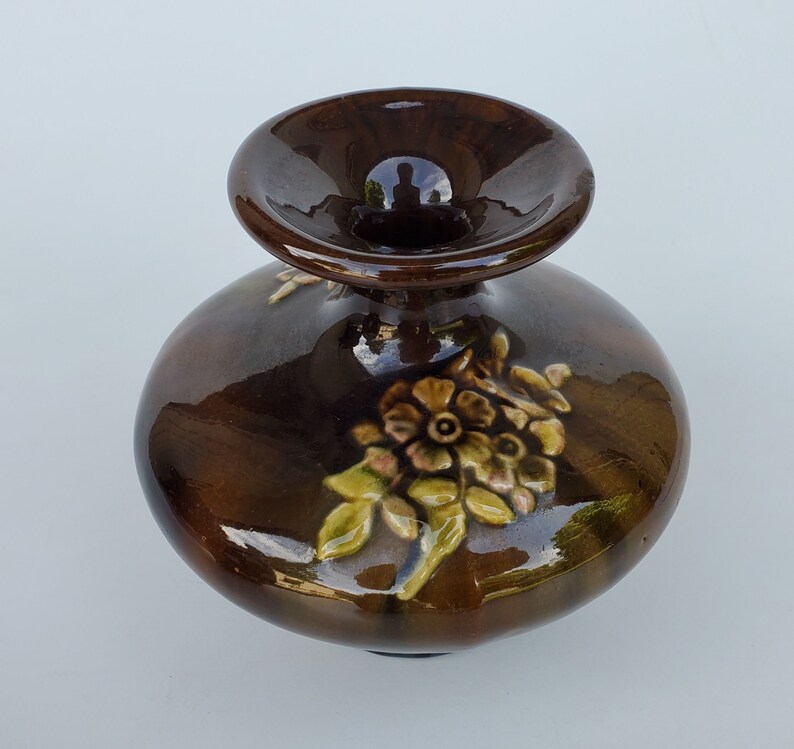 May include: A small brown ceramic vase with a wide opening and a floral design. The vase has a glossy finish and features a raised floral design in shades of brown, yellow, and green.