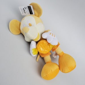 Puede incluir: Un juguete de peluche Mickey Mouse amarillo sosteniendo un caramelo amarillo. El juguete tiene una cara blanca con ojos negros y una nariz negra. El juguete lleva una camisa y pantalones cortos amarillos.