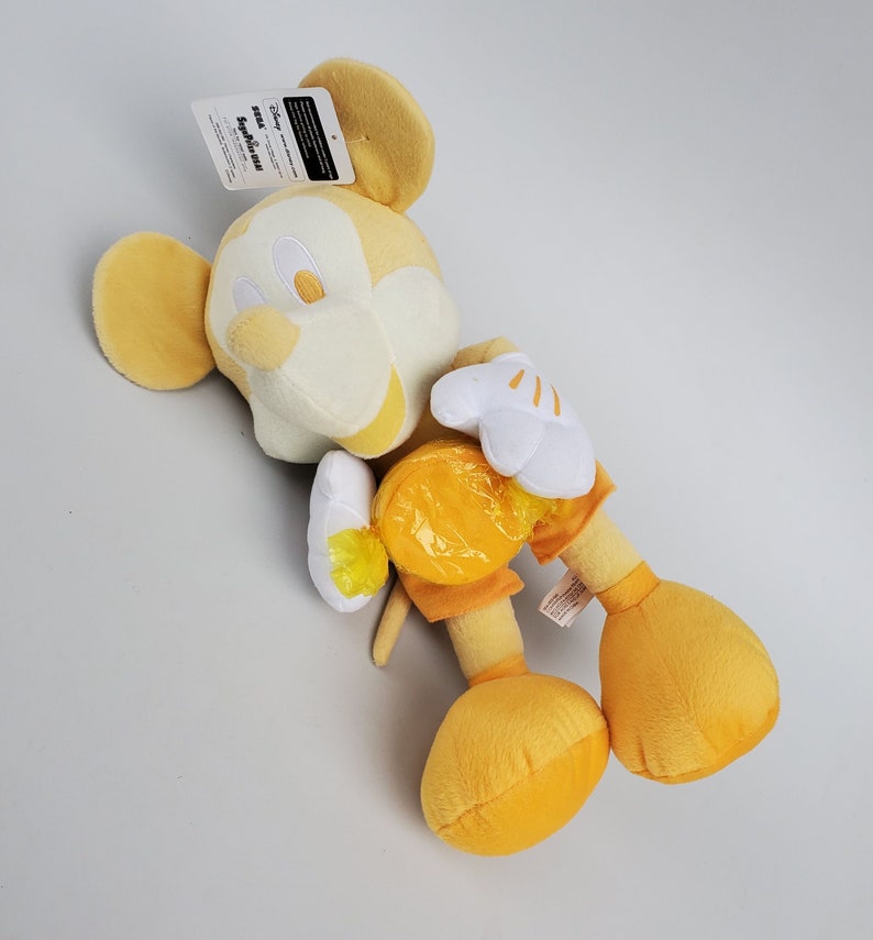 Puede incluir: Un juguete de peluche Mickey Mouse amarillo sosteniendo un caramelo amarillo. El juguete tiene una cara blanca con ojos negros y una nariz negra. El juguete lleva una camisa y pantalones cortos amarillos.