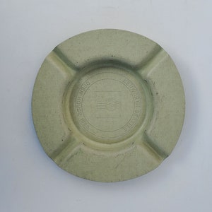 May include: A round, light green ashtray with four cigarette rests. The center features a circular design with the text "OPEN HOUSE 1985" and "KENWORTH SEATTLE". The ashtray has a textured surface.