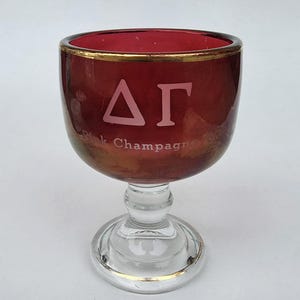 May include: A vintage pink champagne glass with a gold rim and base. The glass features the Greek letters Delta and Gamma in white, along with the words "Pink Champagne '80". The glass has a clear stem and base.