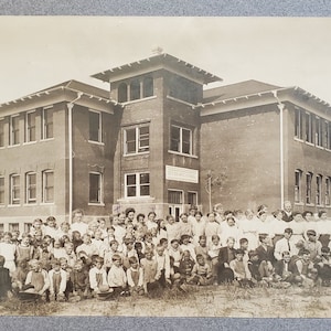 Op de afbeelding: Een zwart-witfoto van een bakstenen schoolgebouw met een grote groep kinderen die ervoor staan. De school is gelabeld met "Entrance School".