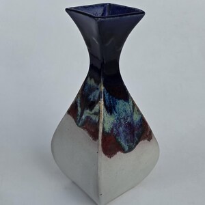 May include: A ceramic vase with a square top and a flared, diamond-shaped body. The top is dark blue, transitioning to a blend of blue, green, and maroon. The base is white, creating a striking contrast.