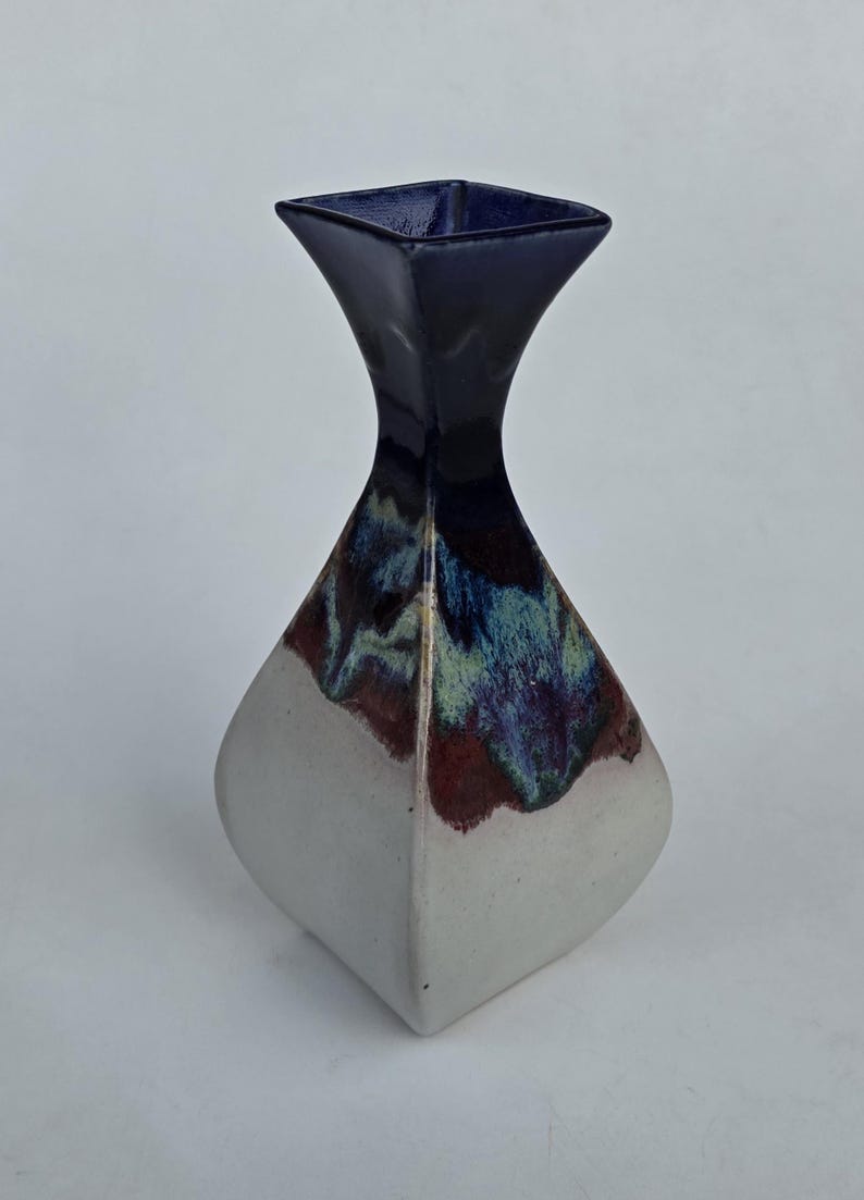 May include: A ceramic vase with a square top and a flared, diamond-shaped body. The top is dark blue, transitioning to a blend of blue, green, and maroon. The base is white, creating a striking contrast.