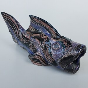 May include: A ceramic fish sculpture with a purple, black, and pink glaze. The fish has a detailed design of swirling lines and a large open mouth.