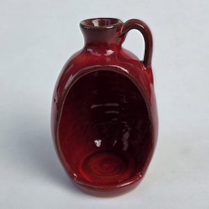 May include: A glossy, deep red ceramic vessel featuring a rounded body with a large, hollowed-out front opening, a narrow neck, and a single curved handle on the side. The interior of the hollow section is smooth and finished in the same red glaze.