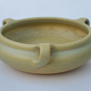 May include: A round, light yellow ceramic bowl with two small handles on the sides and one on the front. The bowl has a wide, shallow design and a slightly textured surface, suitable for decorative or functional use.