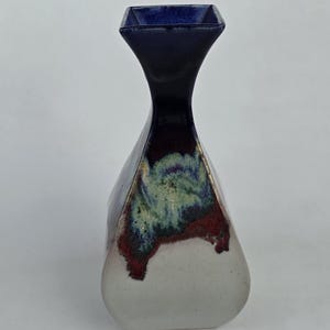 May include: A ceramic vase with a dark blue, square-shaped top and a flared body. The vase features a white base, transitioning to a red and green mottled pattern, and a dark blue neck. The vase has a unique, geometric shape.