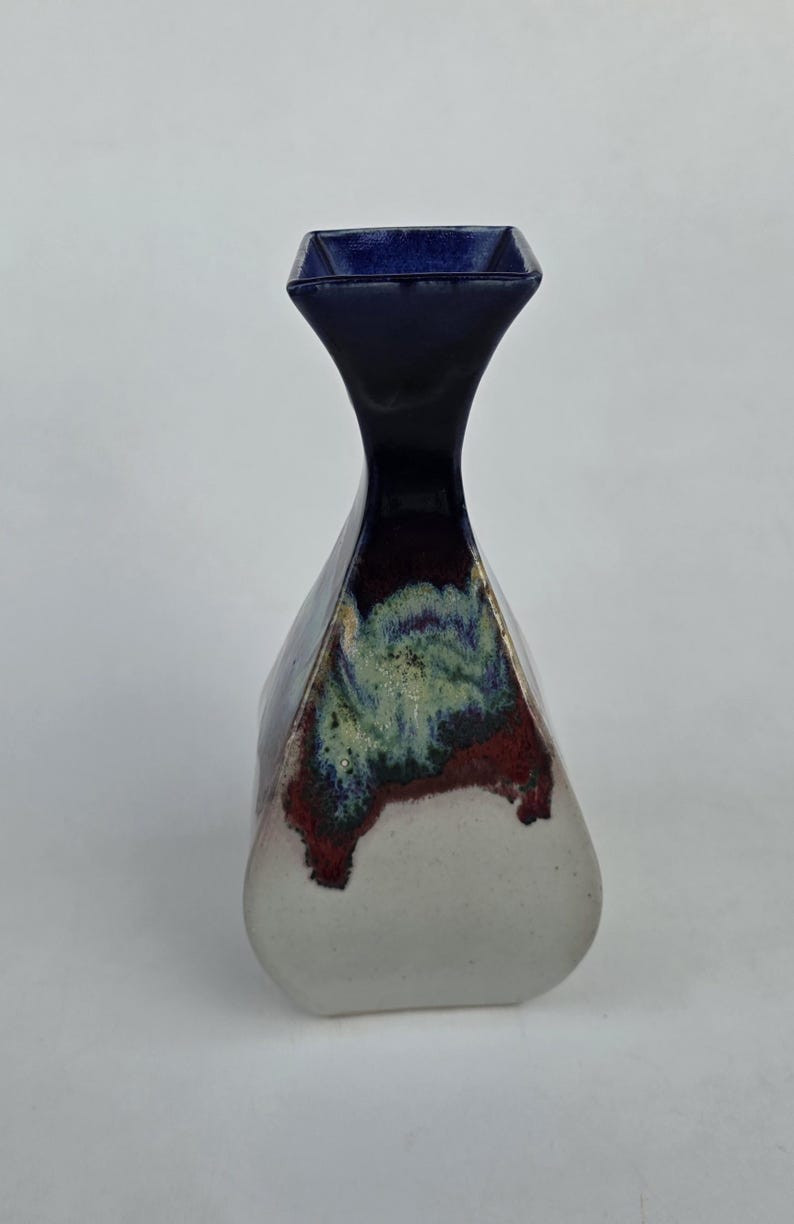 May include: A ceramic vase with a dark blue, square-shaped top and a flared body. The vase features a white base, transitioning to a red and green mottled pattern, and a dark blue neck. The vase has a unique, geometric shape.