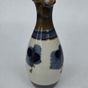 May include: A ceramic carafe with a narrow neck and a flared spout. The body is cream-colored with dark blue, abstract floral-like brushstroke patterns. The neck features a glossy brown and dark blue glaze that transitions into the cream base.