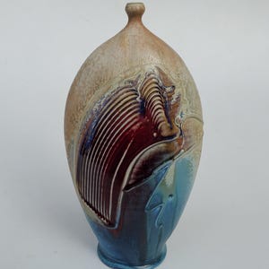 May include: A handcrafted ceramic vase with a unique, organic shape. The vase features a textured surface with a color palette of beige, burgundy, and blue. The design includes a ribbed pattern and abstract shapes, adding visual interest.