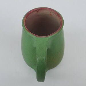 May include: A handmade green ceramic mug with a handle. The interior rim is a reddish-pink color. The mug has a textured surface and a rounded shape, perfect for holding beverages.