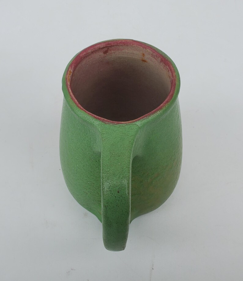 May include: A handmade green ceramic mug with a handle. The interior rim is a reddish-pink color. The mug has a textured surface and a rounded shape, perfect for holding beverages.