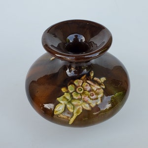 May include: A small, brown ceramic vase with a wide mouth and a floral design. The vase has a glossy finish and features a cluster of pink and yellow flowers with green leaves.