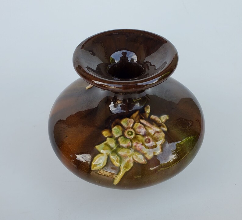 May include: A small, brown ceramic vase with a wide mouth and a floral design. The vase has a glossy finish and features a cluster of pink and yellow flowers with green leaves.