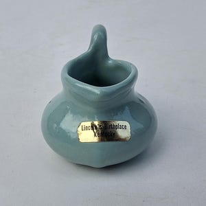 May include: A small, light blue ceramic pitcher with a curved handle. The pitcher has a rounded body and a small opening. A gold label on the front reads "Lincoln's Birthplace Kentucky".