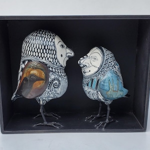May include: Two ceramic birds in a black frame. The bird on the left is white with black and brown details. The bird on the right is white with black, blue, and turquoise details.
