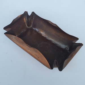 May include: A handcrafted, wooden ashtray with a unique, organic shape. The ashtray is a rich, dark brown color with a smooth, polished finish. It features a rectangular design with curved edges and two notched areas for resting cigarettes.