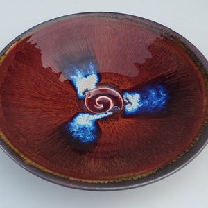 May include: A round ceramic bowl with a glossy finish. The bowl features a deep red-brown color with blue and white accents radiating from the center. A spiral design is at the center of the bowl, and the rim is a darker brown.