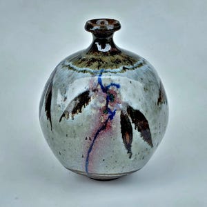 May include: A handcrafted ceramic vase with a round body and a narrow neck. The vase features a speckled, light grey glaze with brown, blue, and red abstract designs. The vase is approximately 18 cm tall.