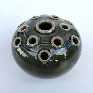 May include: A spherical ceramic flower frog vase with a glossy dark olive green glaze. The top surface features multiple raised circular openings for stems, each rimmed with a lighter beige tone, creating a functional and textured design.
