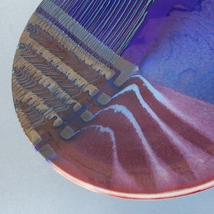 May include: A ceramic plate with a blue, purple, and red glaze. The plate has a unique design with a wavy pattern and a geometric pattern.
