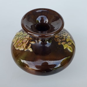May include: A brown ceramic vase with a wide opening and floral designs. The vase has a glossy finish and is decorated with raised flowers in shades of pink, green, and brown.