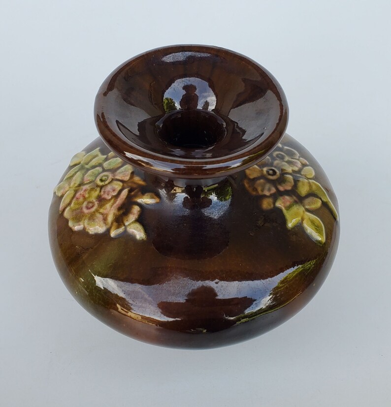 May include: A brown ceramic vase with a wide opening and floral designs. The vase has a glossy finish and is decorated with raised flowers in shades of pink, green, and brown.